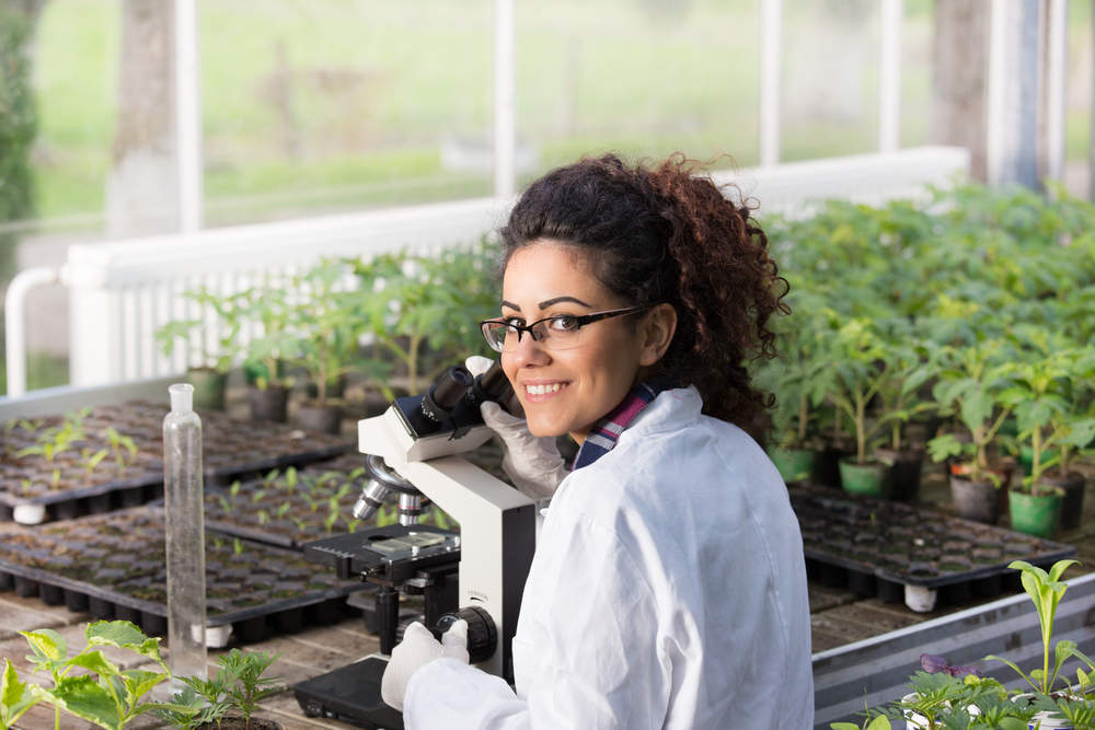 Young,Biologist,Sitting,At,Microscope,With,Seedlings,Around,Her,In